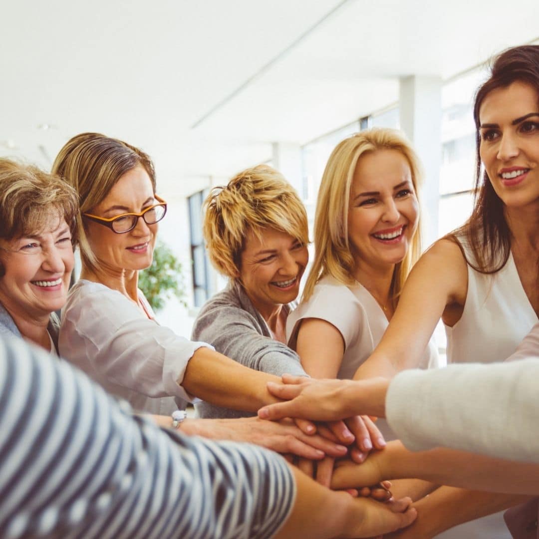 Grupo de mulheres coroas reunidas e sorrindo, com as mãos uma sobre a outra, mantendo o foco na união e no sucesso compartilhado.