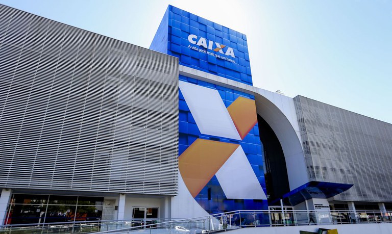 Minha Casa Minha Vida 2026 em frente à fachada da agência central da Caixa Econômica Federal, com destaque para a logomarca azul e arquitetura moderna.