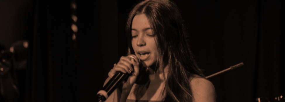 Mulher cantando com microfone em frente a banner com logomarcas da Fames, Fapes e parceiros do Projeto Música para Todos.