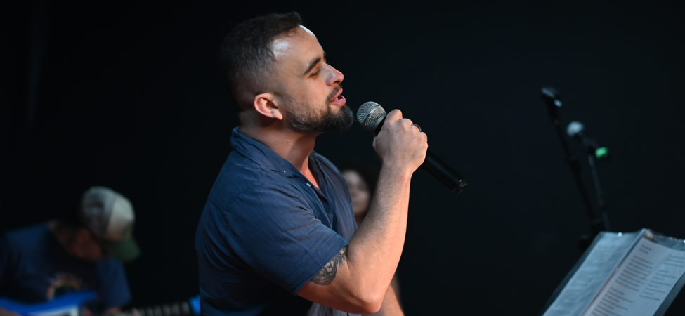 Cantor de lado no palco segurando microfone em apresentação musical com fundo fosco.