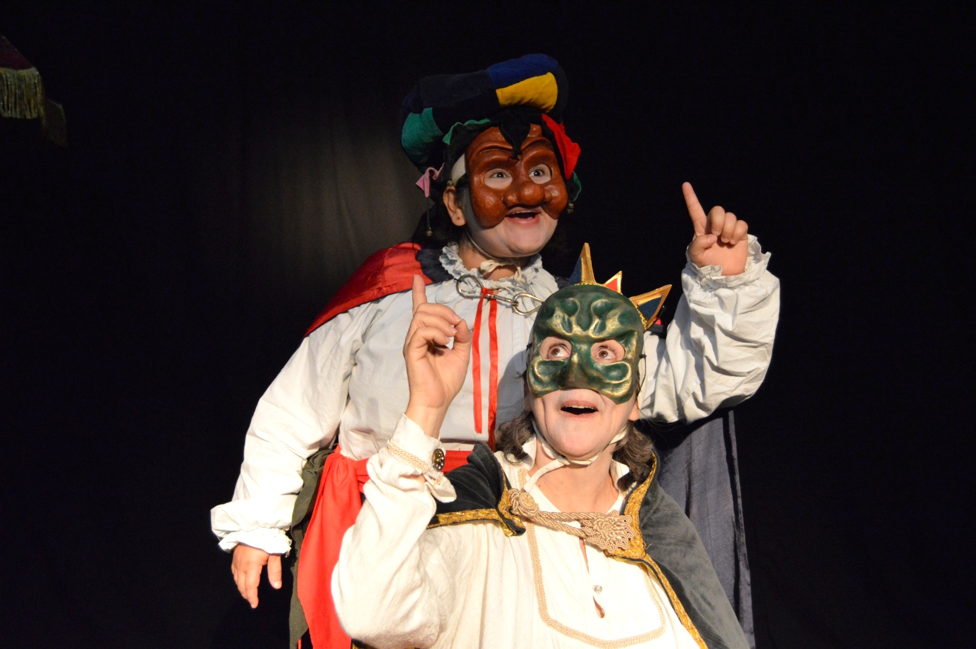 Fotografia em plano médio de dois artistas de teatro em pé, lado a lado, usando máscaras teatrais expressivas cobrindo o rosto inteiro.