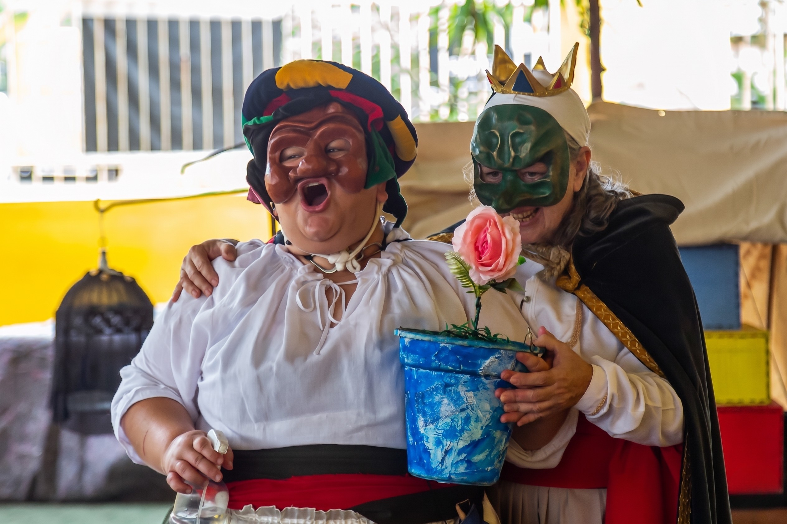 Atrizes de teatro em palco escolar com máscaras e figurinos coloridos.