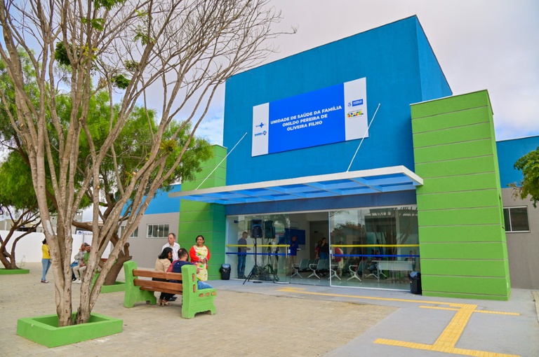 Fachada da Unidade de Saúde da Família com pessoas aguardando atendimento em bancos externos e internos, ilustrando a necessidade de ampliação da Saúde no Espírito Santo.c