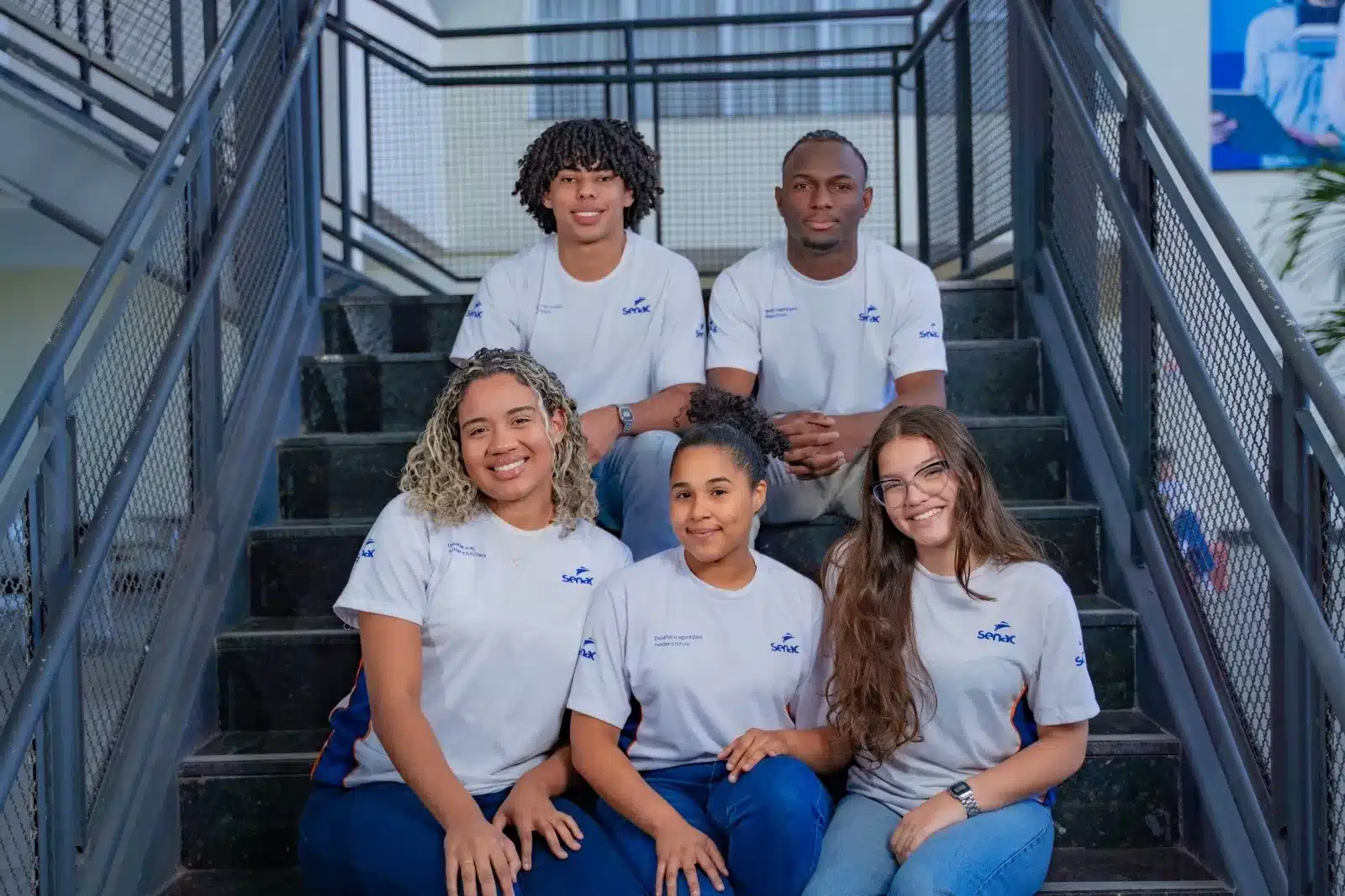 Grupo de jovens estudantes uniformizados do Senac Espírito Santo, sorrindo e sentados nos degraus da instituição, representando a união e a educação profissional.