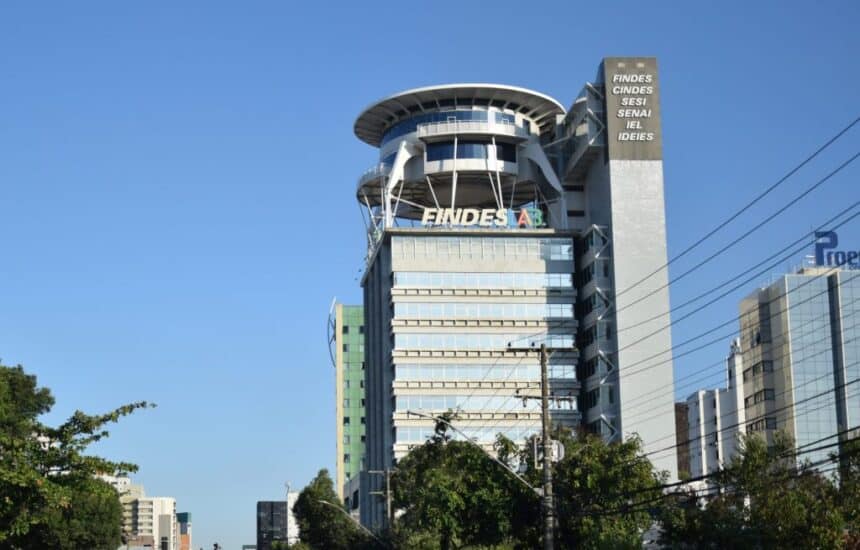"Fotografia panorâmica tirada de baixo para cima (ângulo contra-plongée) que mostra toda a imponente fachada de vidro e estrutura do edifício sede da FINDES (Federação das Indústrias do Espírito Santo), em Vitória. A imagem destaca a entrada principal e a verticalidade do prédio contra o céu, onde será realizada a Rodada de Crédito."