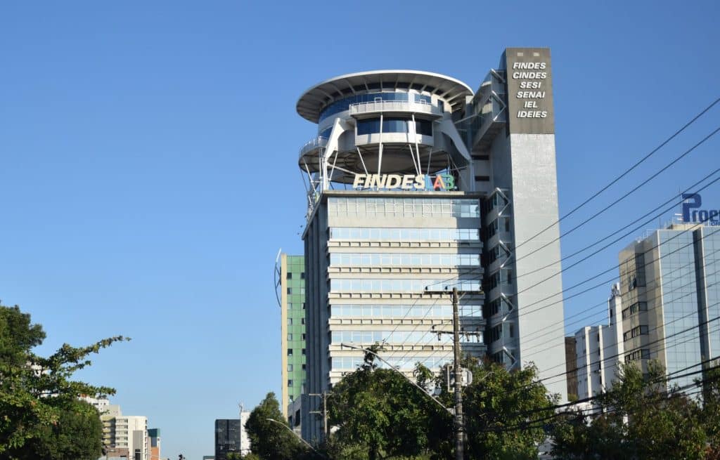 "Fotografia panorâmica tirada de baixo para cima (ângulo contra-plongée) que mostra toda a imponente fachada de vidro e estrutura do edifício sede da FINDES (Federação das Indústrias do Espírito Santo), em Vitória. A imagem destaca a entrada principal e a verticalidade do prédio contra o céu, onde será realizada a Rodada de Crédito."