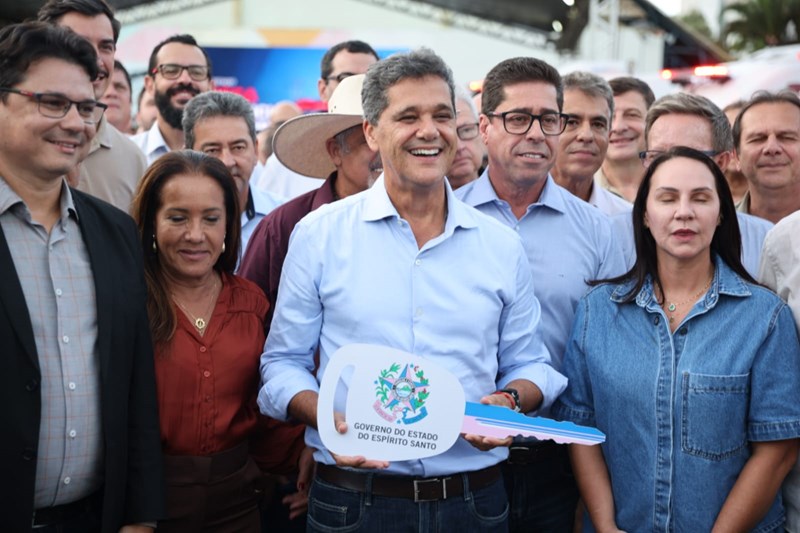 Governador Ricardo Ferraço sorrindo e segurando uma chave simbólica, cercado por diversas pessoas, com a frota de novas ambulâncias ao fundo.