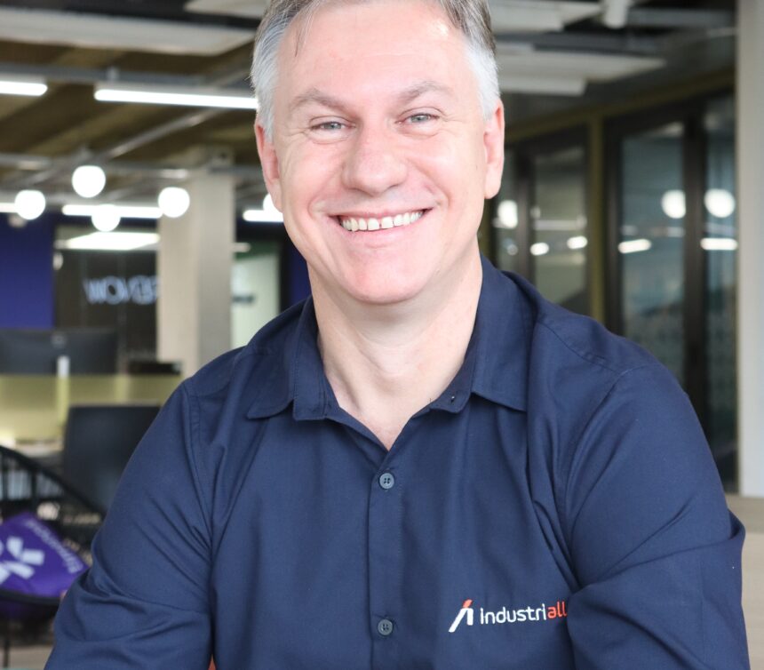 Retrato de Rodrigo Dal Moro, homem branco de cabelos grisalhos e camisa de uniforme da IndustriALL, sorrindo com o pátio de uma indústria desfocado ao fundo para ilustrar o artigo sobre inteligência artificial.