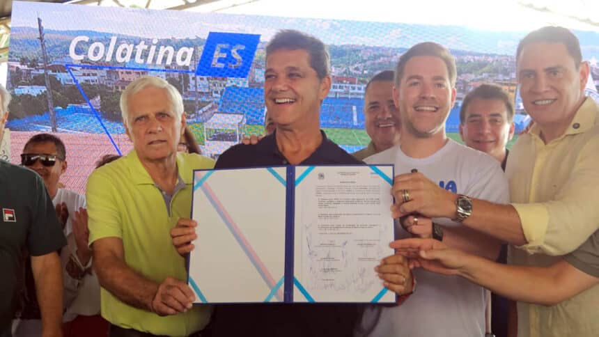Ricardo Ferraço segura documento assinado e sorri ao lado de secretários e autoridades durante evento de anúncio de investimentos em Colatina.