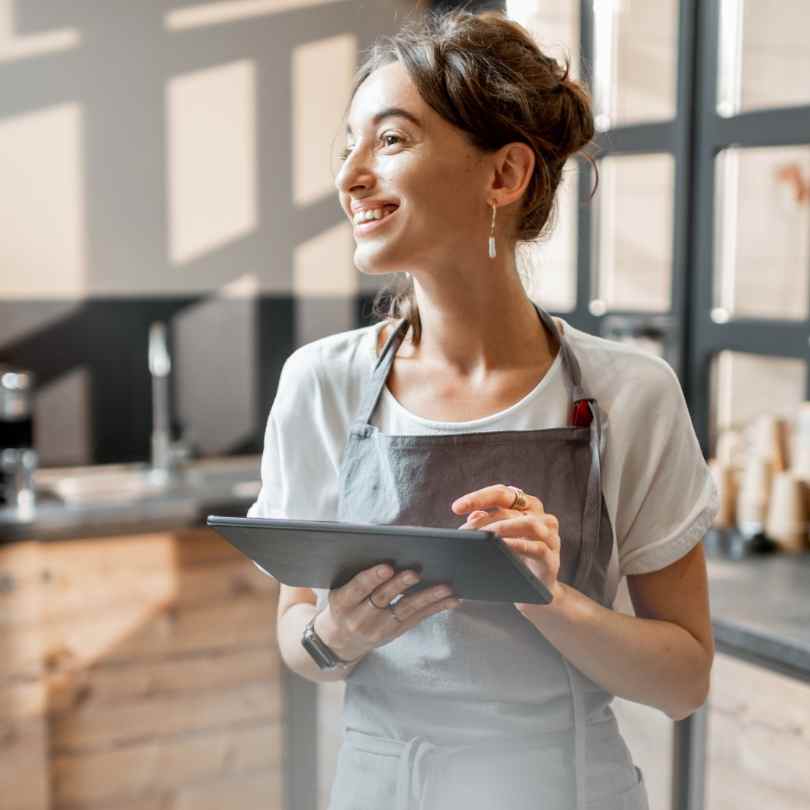 Mulher de aparência simples e trabalhadora, sentada à mesa de casa, sorrindo ao conferir a data de pagamento do Bolsa Família Maio 2026 em um tablet.