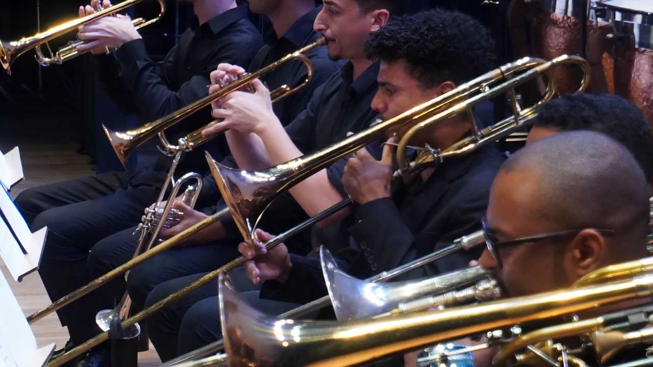 Instrumentistas de sopro posicionados no palco durante a execução de um concerto sinfônico especial em Vitória.