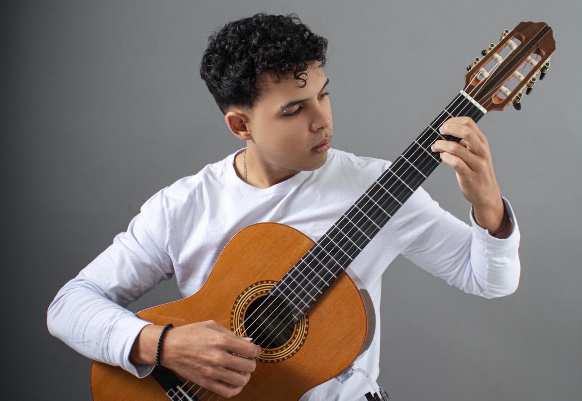 Músico concentrado tocando violão clássico durante apresentação de música instrumental no palco de um teatro antigo.