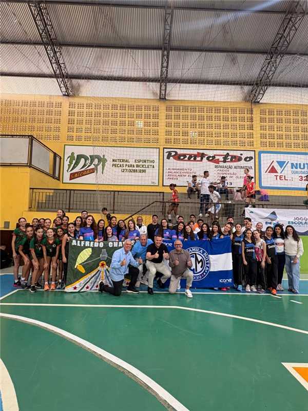 Grupo de meninas adolescentes com uniforme esportivo comemorando vitória em quadra de handebol durante os Jogos Escolares.