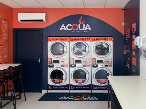 Vista interna de uma lavanderia moderna e automatizada com fileiras de máquinas de lavar e secar de inox brilhantes em ambiente iluminado.