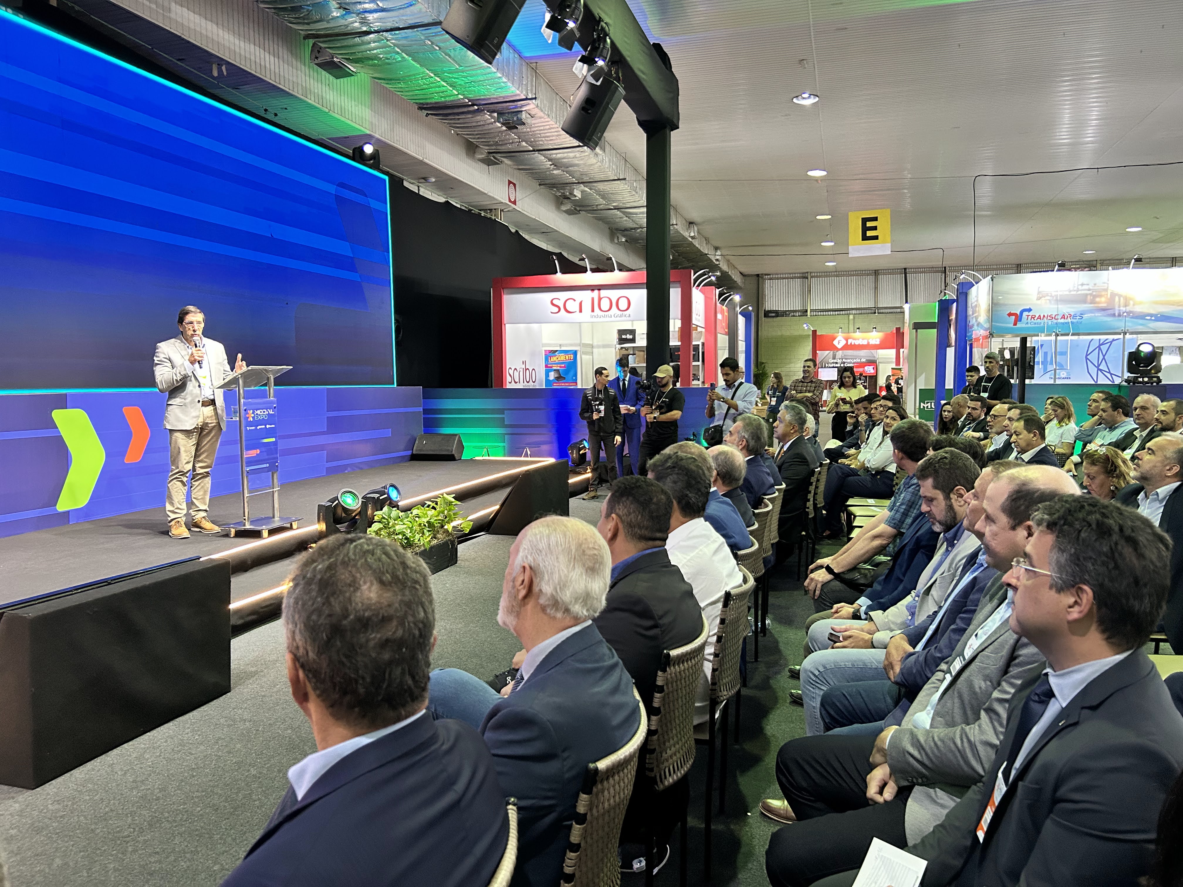 O presidente da Findes, Paulo Baraona, gesticula e fala ao microfone no palco de um evento empresarial, usando terno escuro.