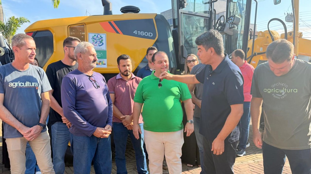 Motoniveladora patrol amarela nova estacionada em frente ao prédio administrativo municipal durante o dia.