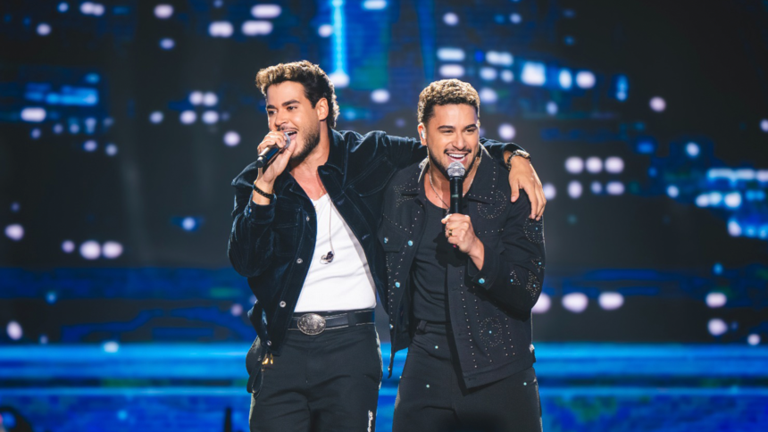 Dupla de cantores do gênero sertanejo se apresentando em um palco iluminado com público ao fundo.