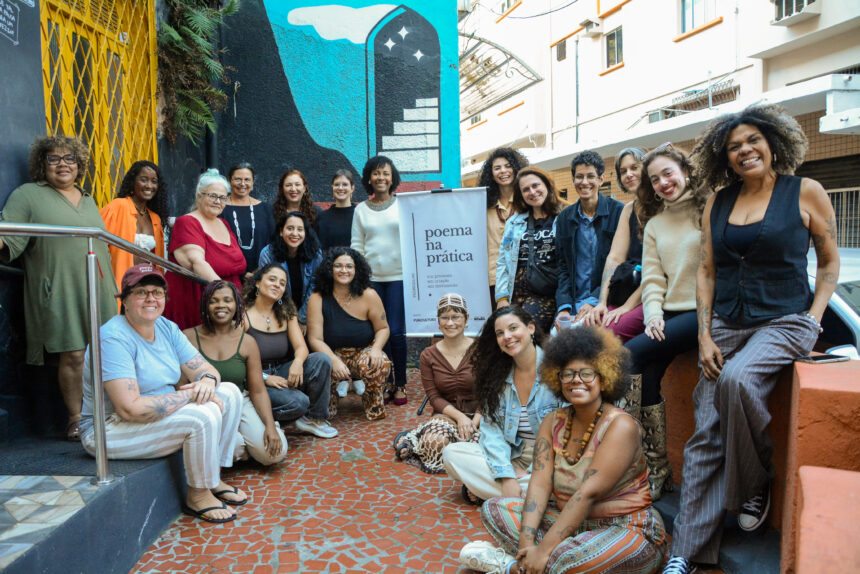 Mãos folheando o novo livro Poema na Prática em uma mesa de madeira durante lançamento cultural.