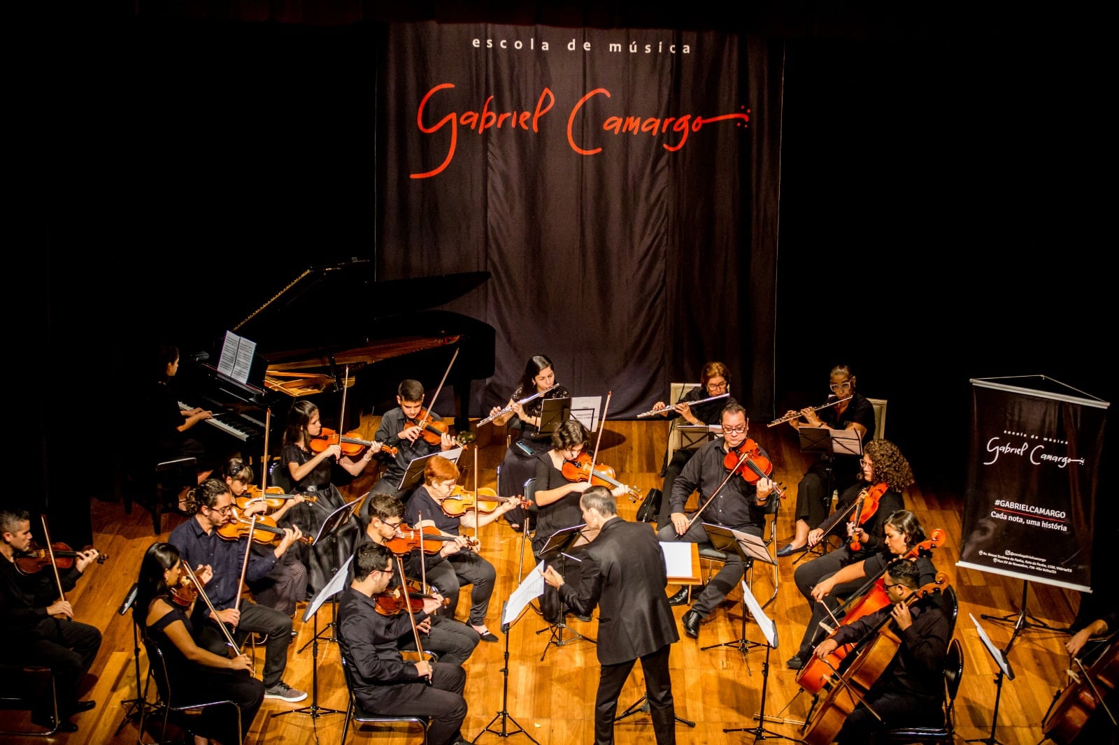 Músicos da Escola Gabriel Camargo com violinos no palco e maestro regendo onde a Casa da Música Sônia Cabral anuncia programação pós-feriado.
