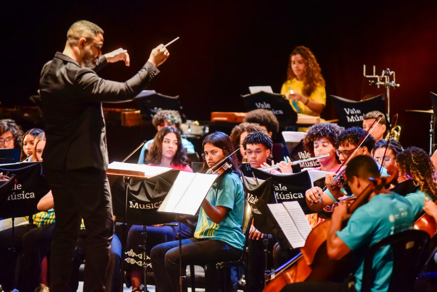 Integrantes da Orquestra Jovem Vale Música apresentando-se no palco com violinos e instrumentos de orquestra.