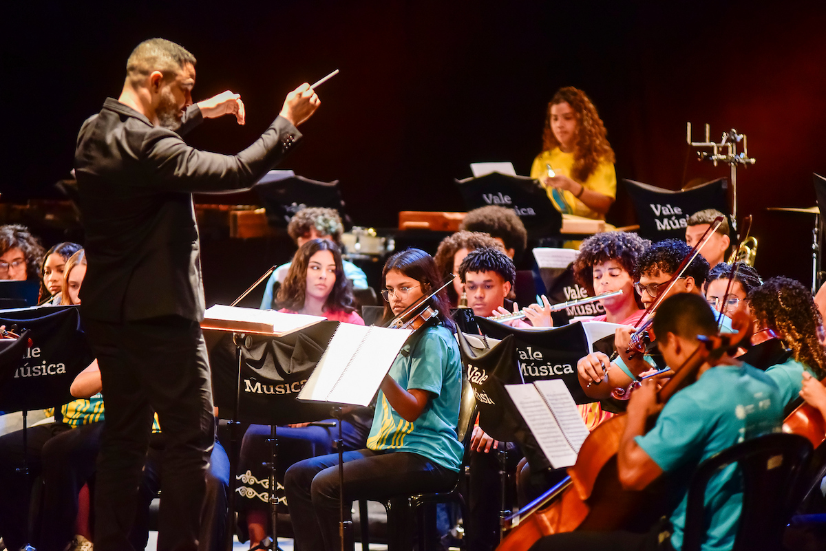 Integrantes da Orquestra Jovem Vale Música apresentando-se no palco com violinos e instrumentos de orquestra.